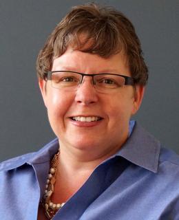 Photograph of a woman with short brown hair, wearing glasses, a blue shirt and a string of pearls.
