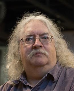 Photograph of a man with long curly light-coloured hair and moustache, wearing glasses and a purple shirt